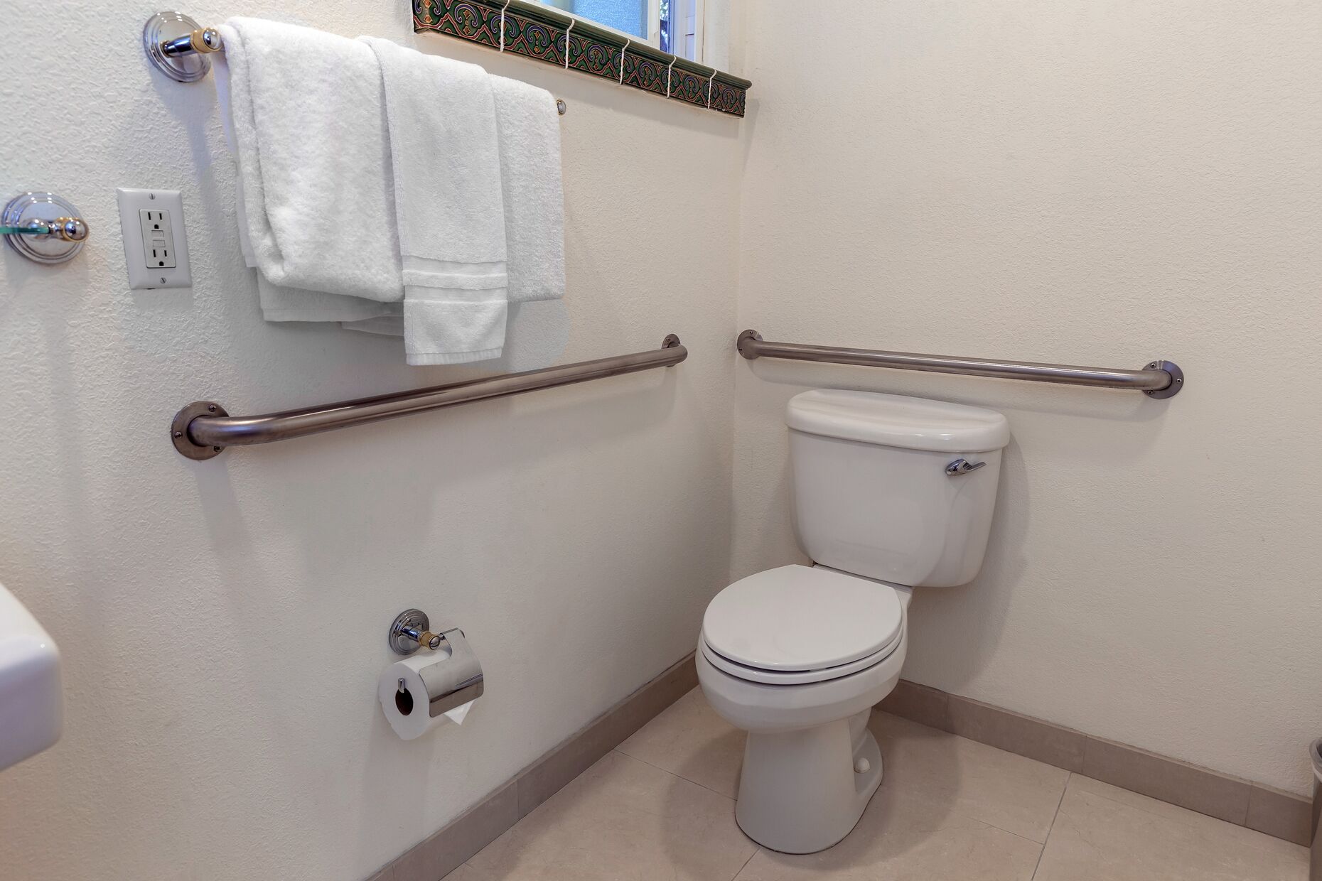 A bathroom with a toilet, two handrails, a towel rack with two towels, a toilet paper holder, and a decorative tile border above the towel rack.