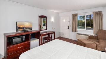 Ein Hotelzimmer mit einem Bett, einem Schreibtisch mit Computer, einem Fernseher, einem Sessel und einem Fenster mit Vorhängen.