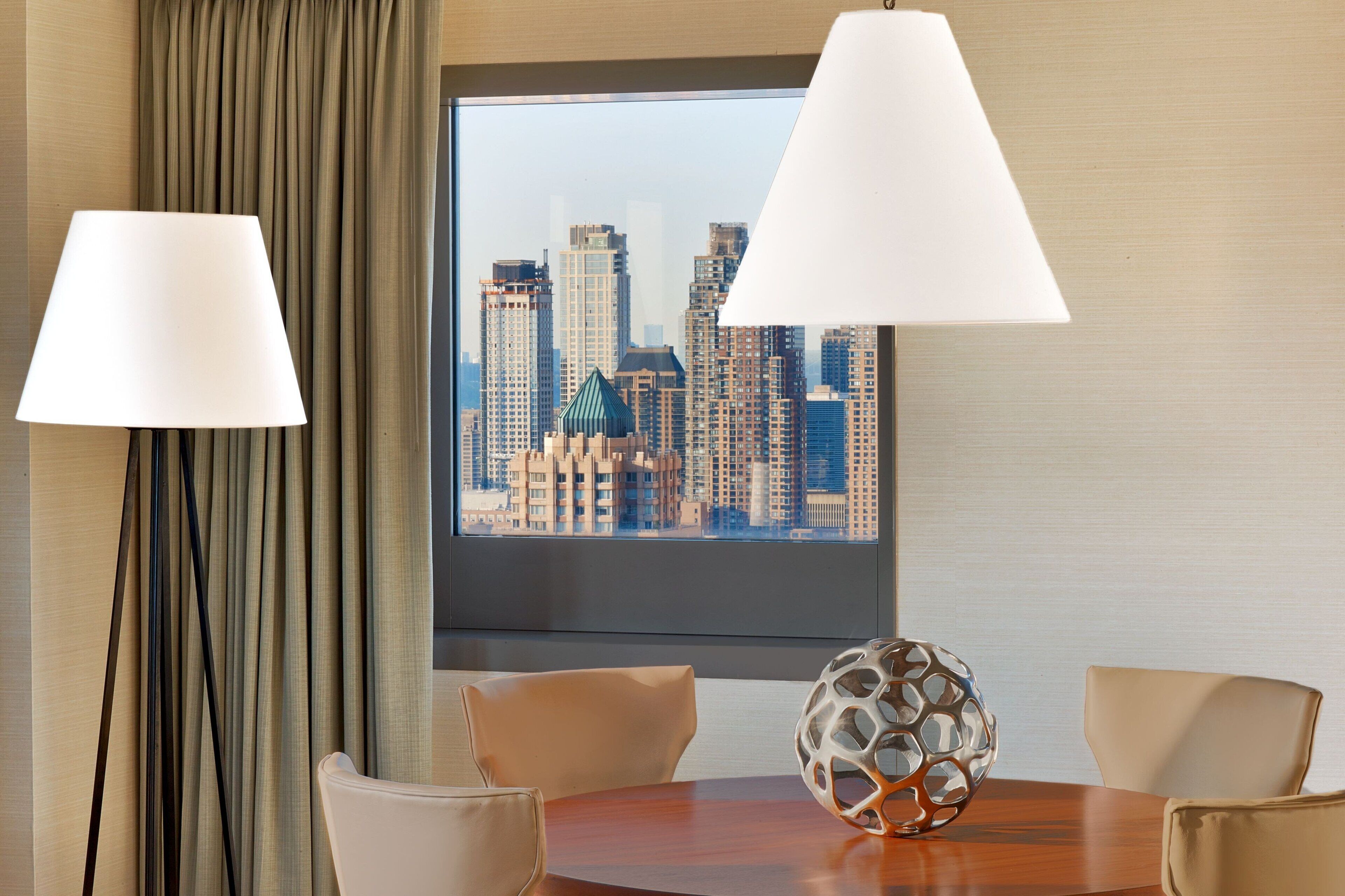 A hotel room with a city view, a round table, two chairs, a decorative vase, and two lamps.