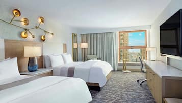 A hotel room with two beds, a desk, a chair, a lamp, and a view of the city through a window.