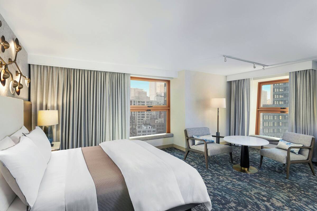 Een hotelkamer met een bed, twee stoelen, een klein tafeltje, uitzicht op de stad en een lamp.