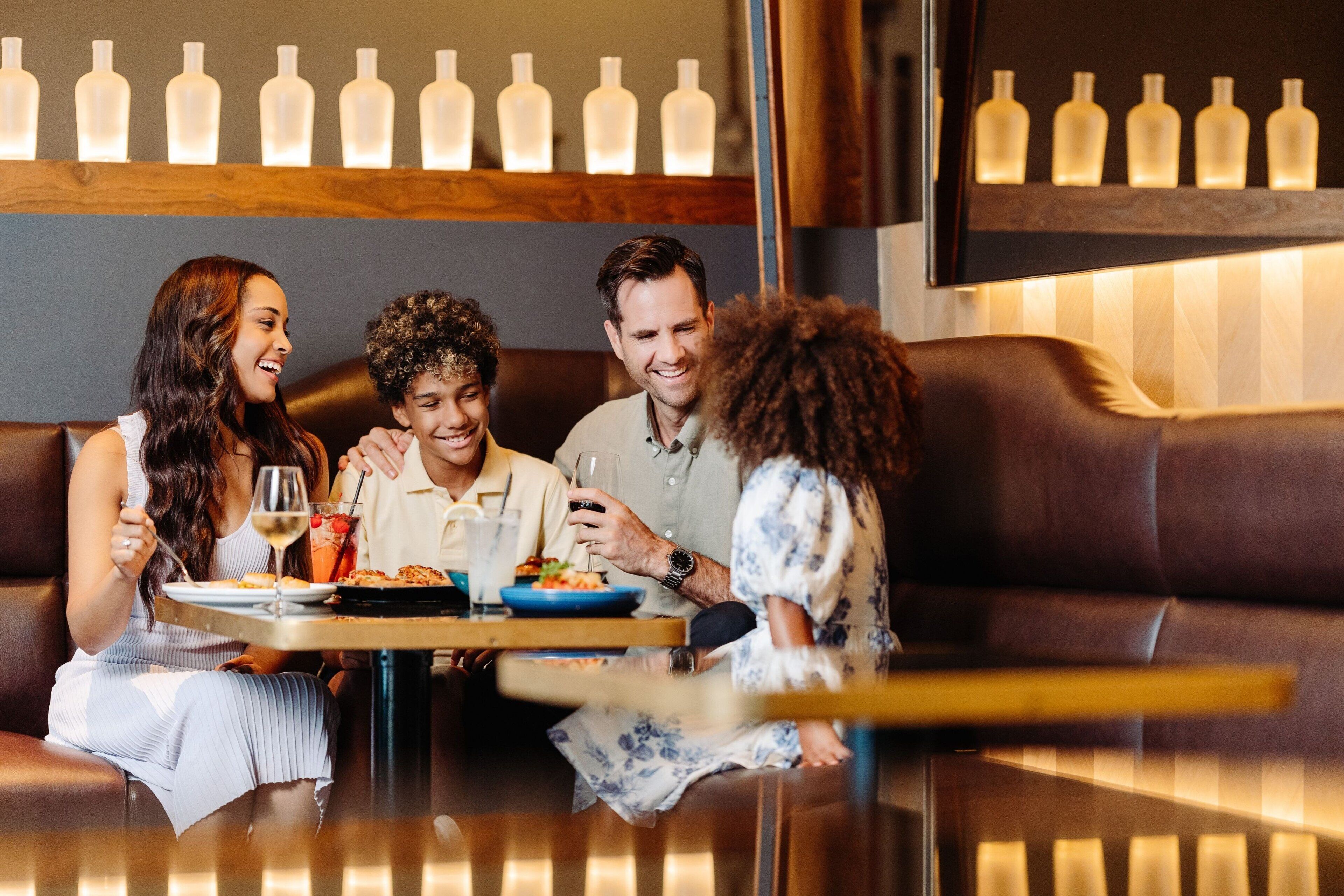 Un grupo de personas cenando en una mesa en un ambiente interior acogedor y bien iluminado, con techo de madera y una fila de botellas en la pared detrás.