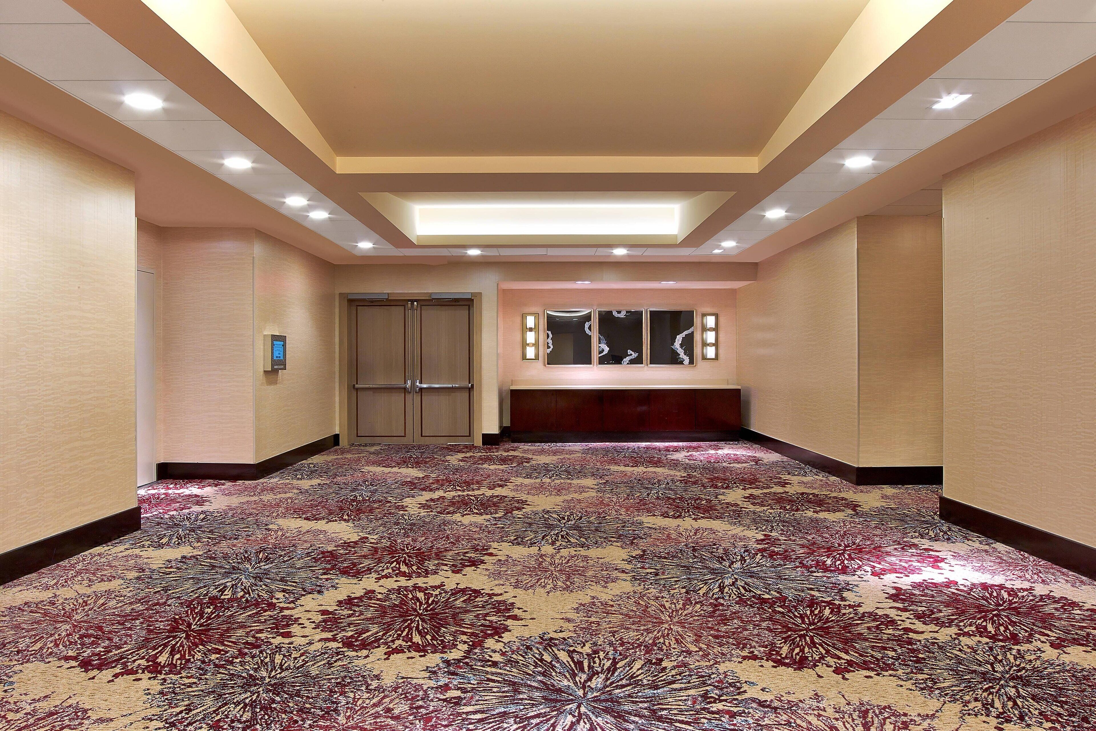 A hotel corridor with a patterned carpet, recessed lighting, a reception desk, and a door.