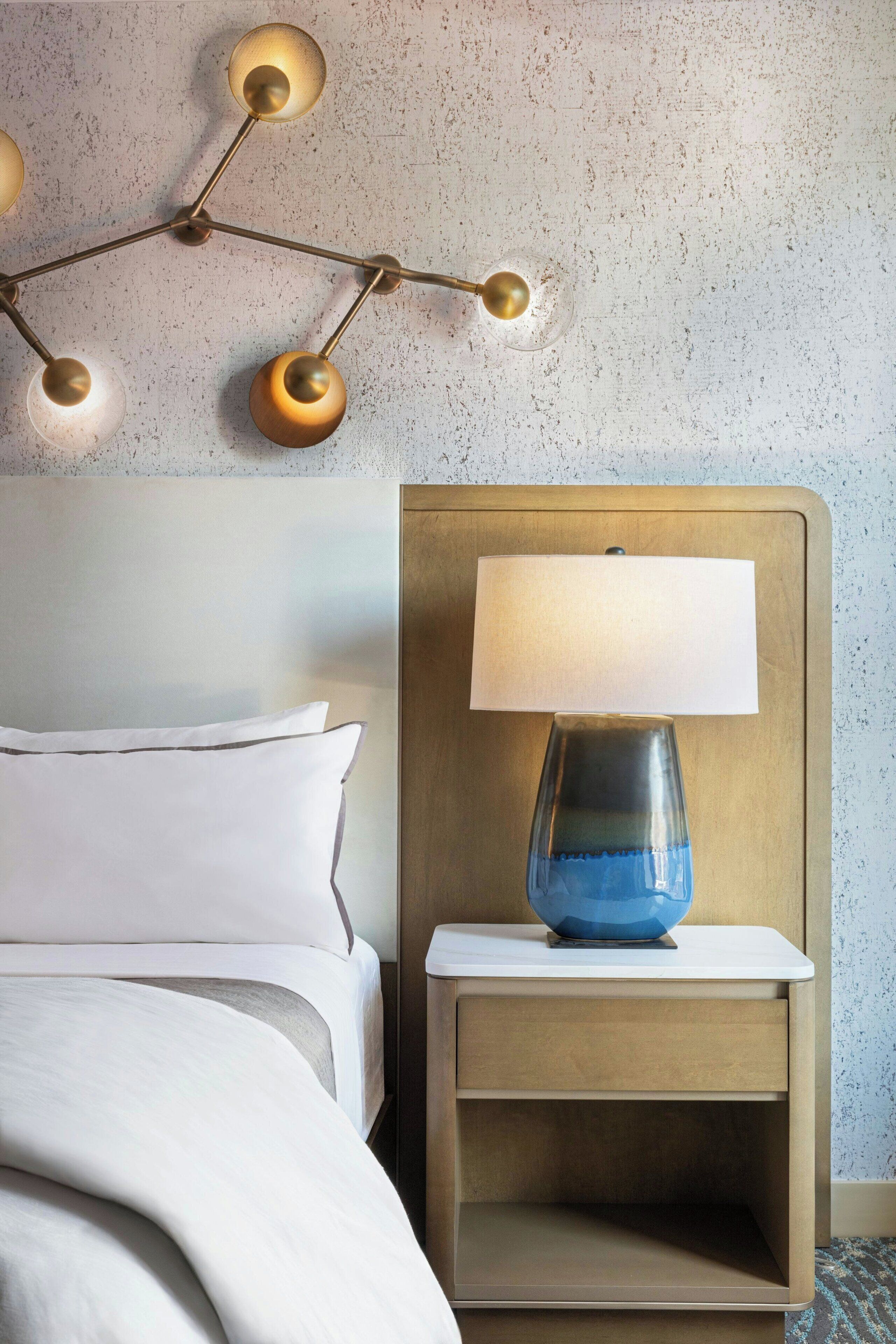 Una cama bien hecha con sábanas blancas, una mesita de noche con lámpara y una lámpara de pared.