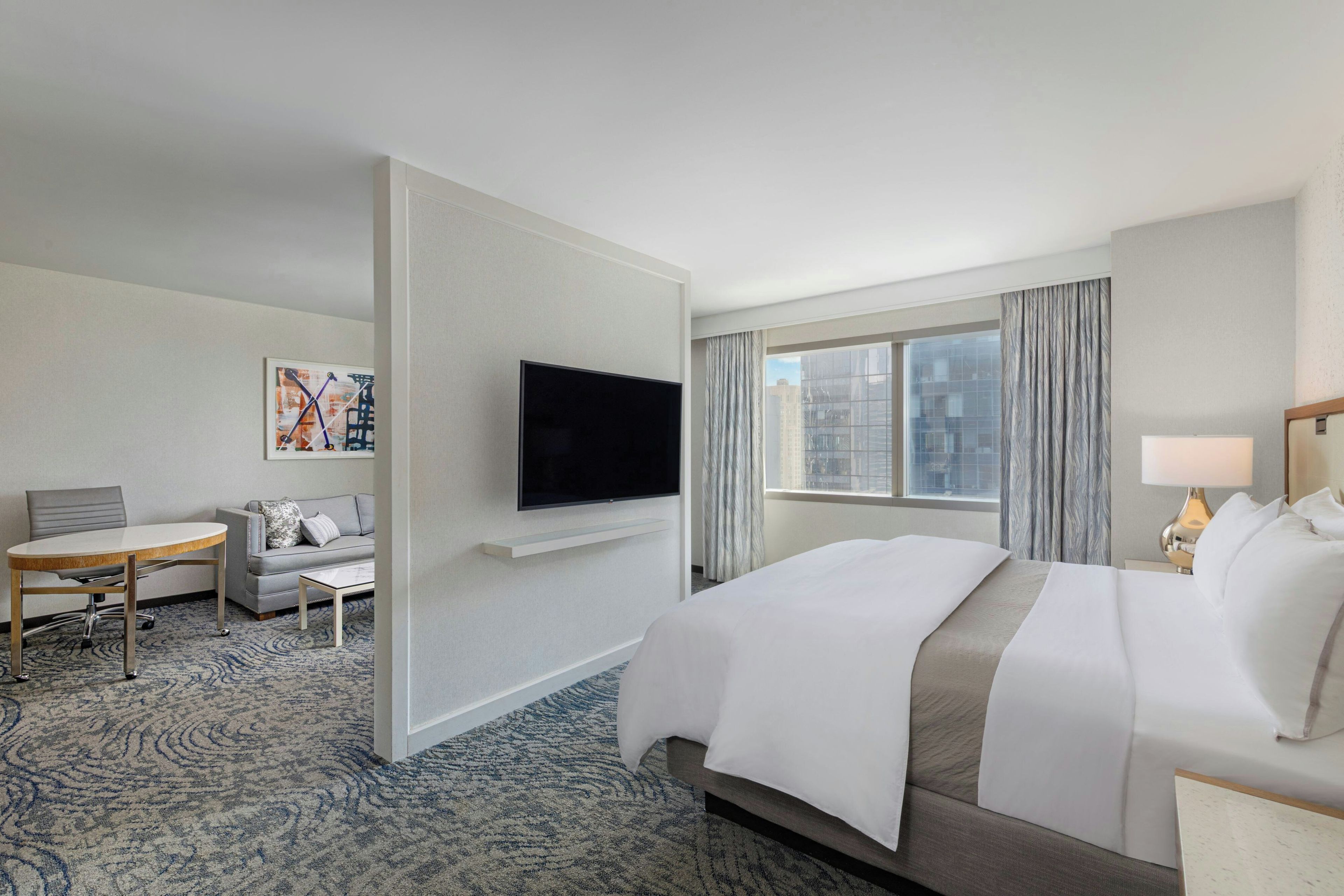 Habitación de hotel con cama, televisión montada en la pared, escritorio con silla y ventana con cortinas.