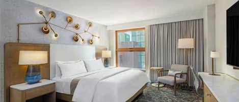 Habitación de hotel con una cama grande, mesitas de noche, un escritorio, una silla, un televisor y una ventana con cortinas.