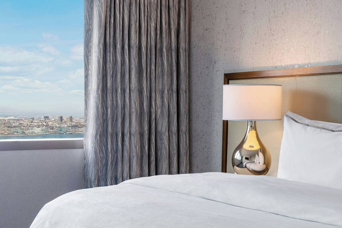 Een hotelkamer met een bed, een lamp, uitzicht op de stad en een gordijn.