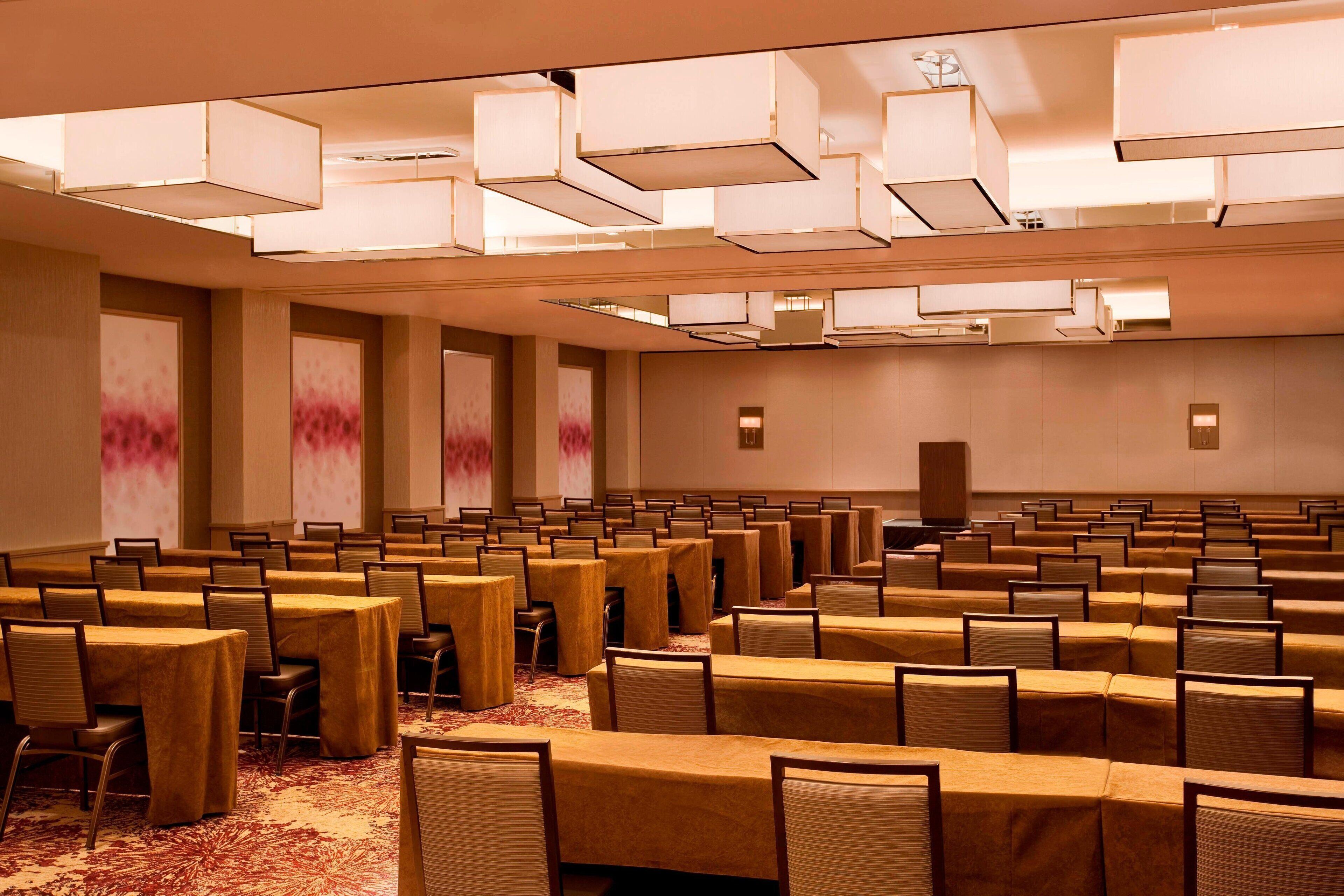 A conference room with rows of tables and chairs, large ceiling lights, and a carpeted floor.