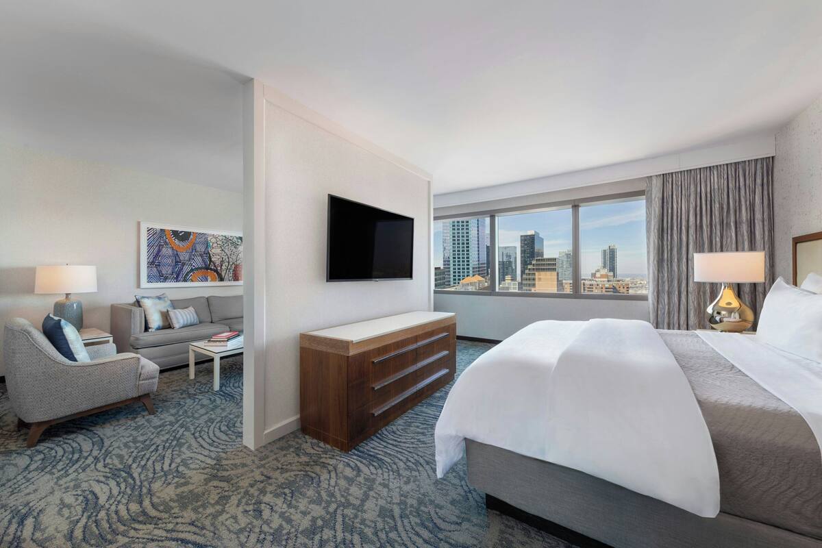 Een hotelkamer met een bed, een bank, een televisie, een bureau en een groot raam met uitzicht op de stad.