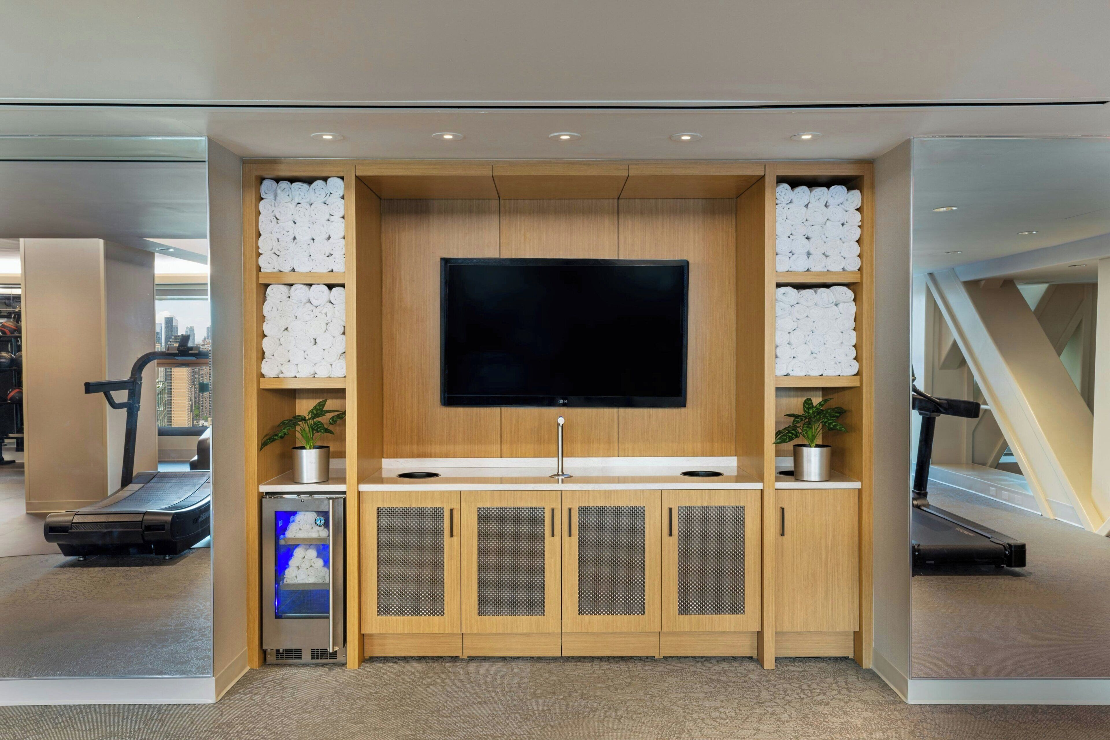 A modern living room with a flat-screen TV, wooden cabinets, a treadmill, and a small refrigerator.