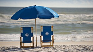 Two beach chairs under a blue umbrella on a sandy beach with the ocean in the background. - SpringHill Suites Pensacola Beach (Pensacola Beach)