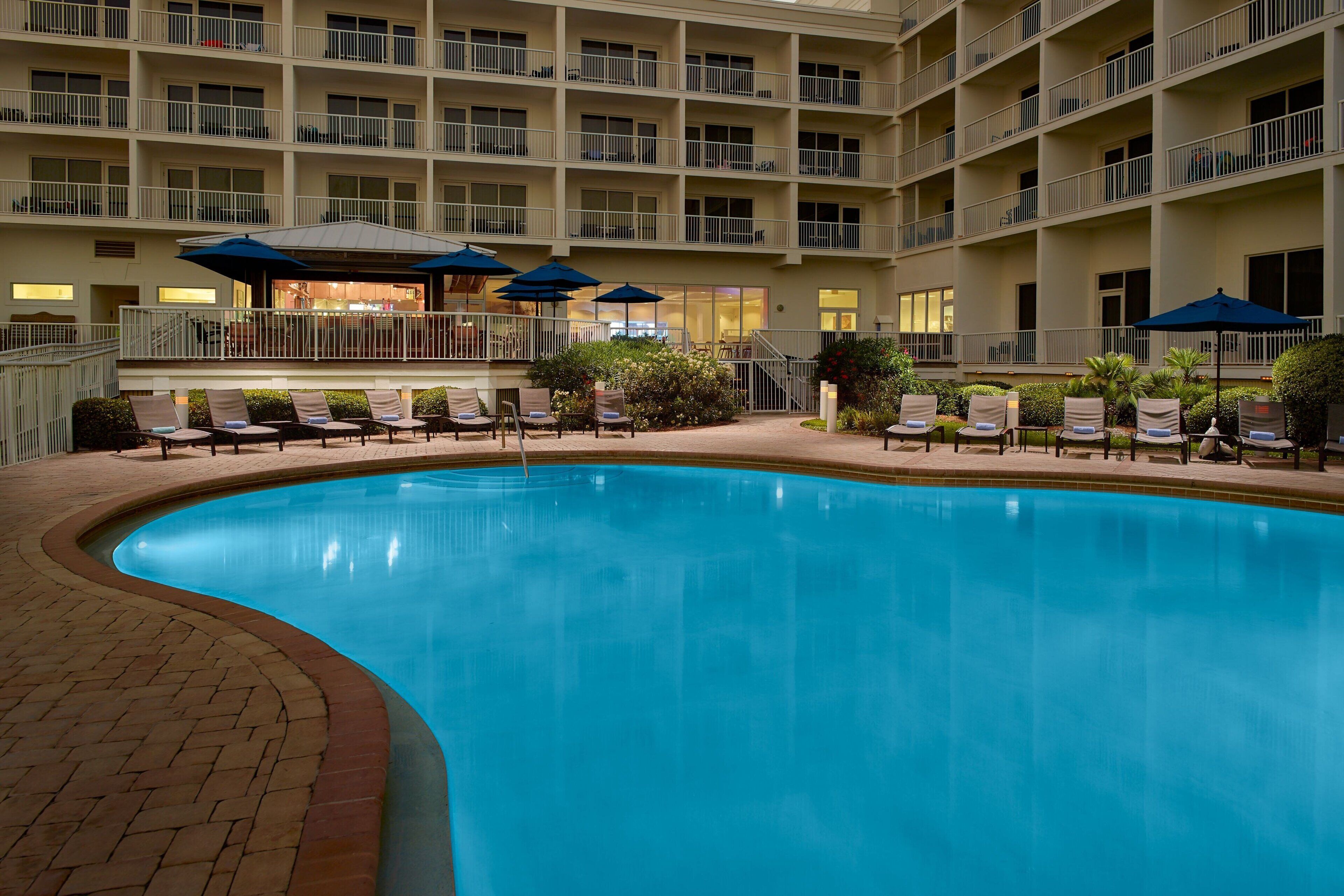 A hotel pool area with lounge chairs and umbrellas, surrounded by a multi-story building with balconies.