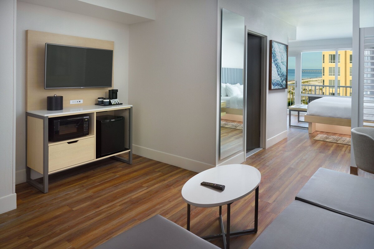 A modern hotel room with a flat-screen TV, a microwave, a small table, and a bed with a view of the beach and buildings.
