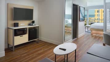 Een moderne hotelkamer met een flatscreen-tv, een magnetron, een klein tafeltje en een bed met uitzicht op het strand en de gebouwen.