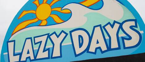 A sign with the text "LAZY DAYS" and "please be seated surf conditions" against a blue background with a sun and clouds illustration.
