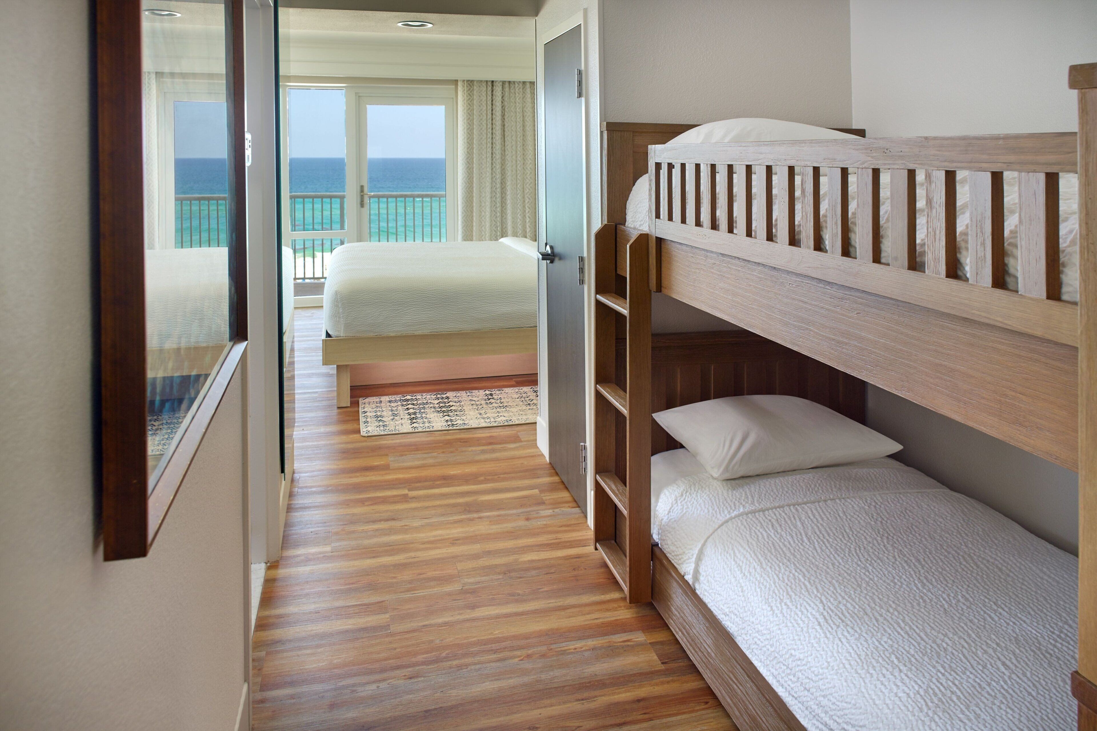 Una stanza con un letto a castello in legno, un letto singolo con materasso bianco e vista sull'oceano attraverso una porta finestra.