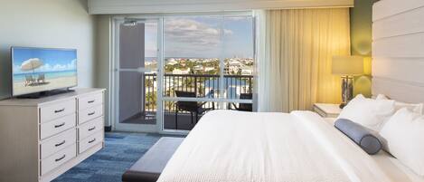 A hotel room with a bed, a TV on a dresser, a balcony with a view of the city, and a lamp on the nightstand.