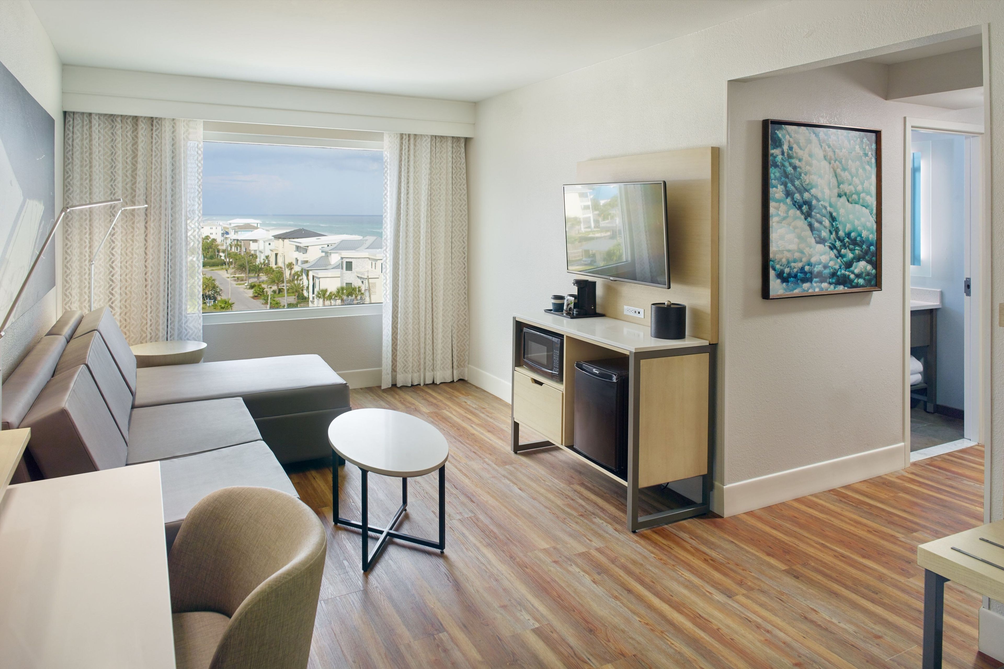 A modern hotel room with a sofa, a small round table, a television, a microwave, and a refrigerator. There is a large window offering a view of the ocean and buildings.