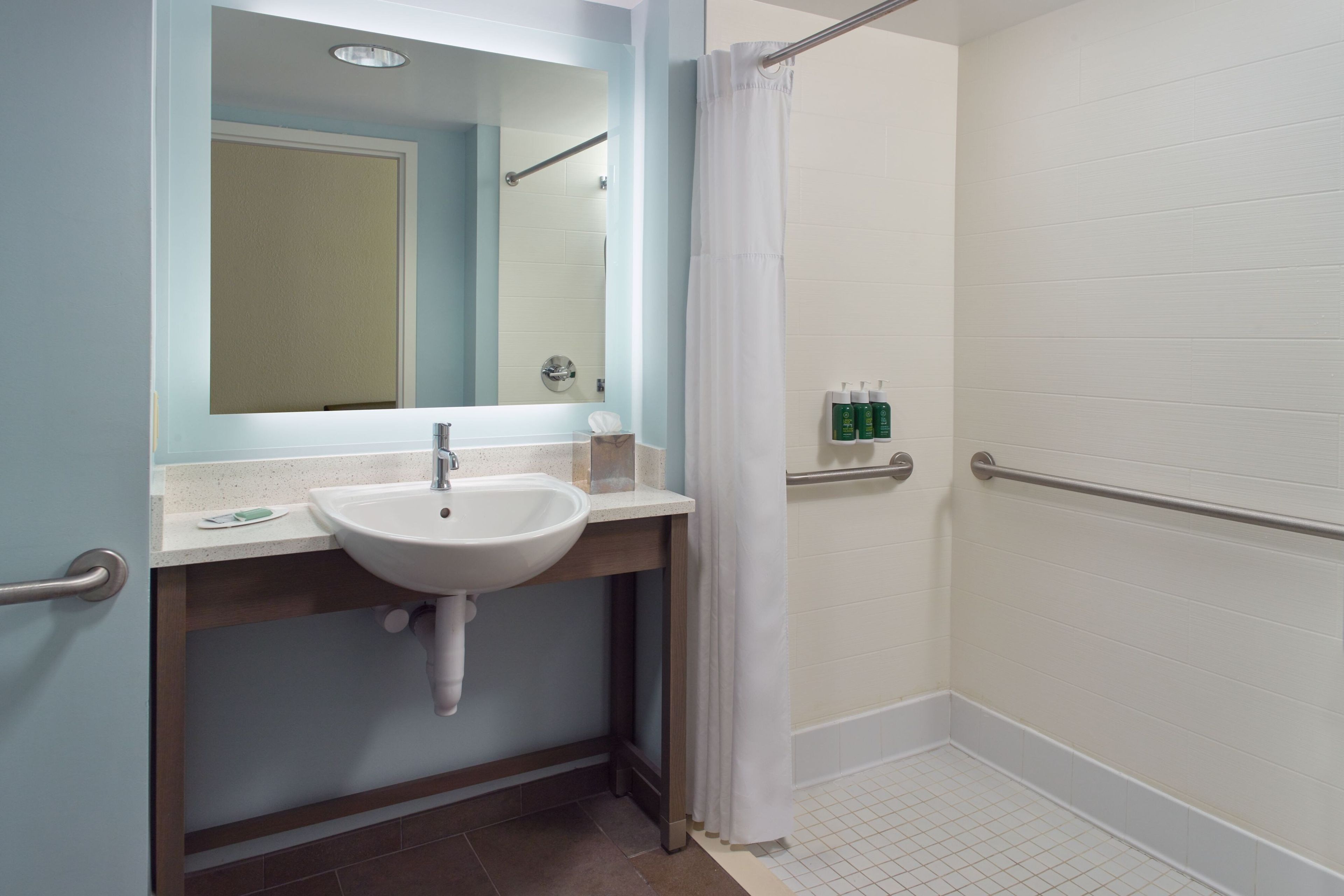 A bathroom with a white sink, a mirror, a shower with a white curtain, and a handrail.