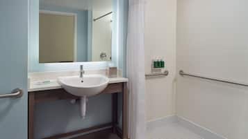 Une salle de bain avec un lavabo blanc, un miroir, une douche avec un rideau blanc et une barre d’appui.