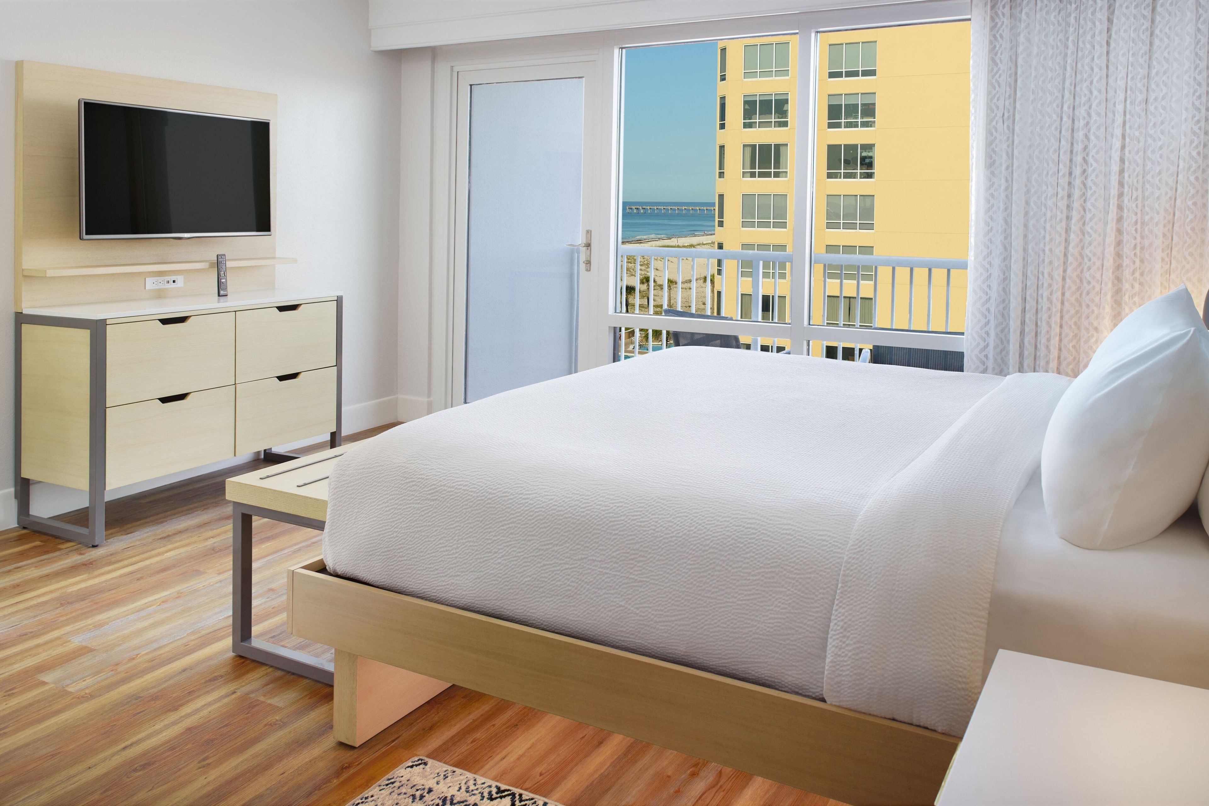 A hotel room with a bed, a TV on a stand, a desk, and a view of a building and beach through the window.