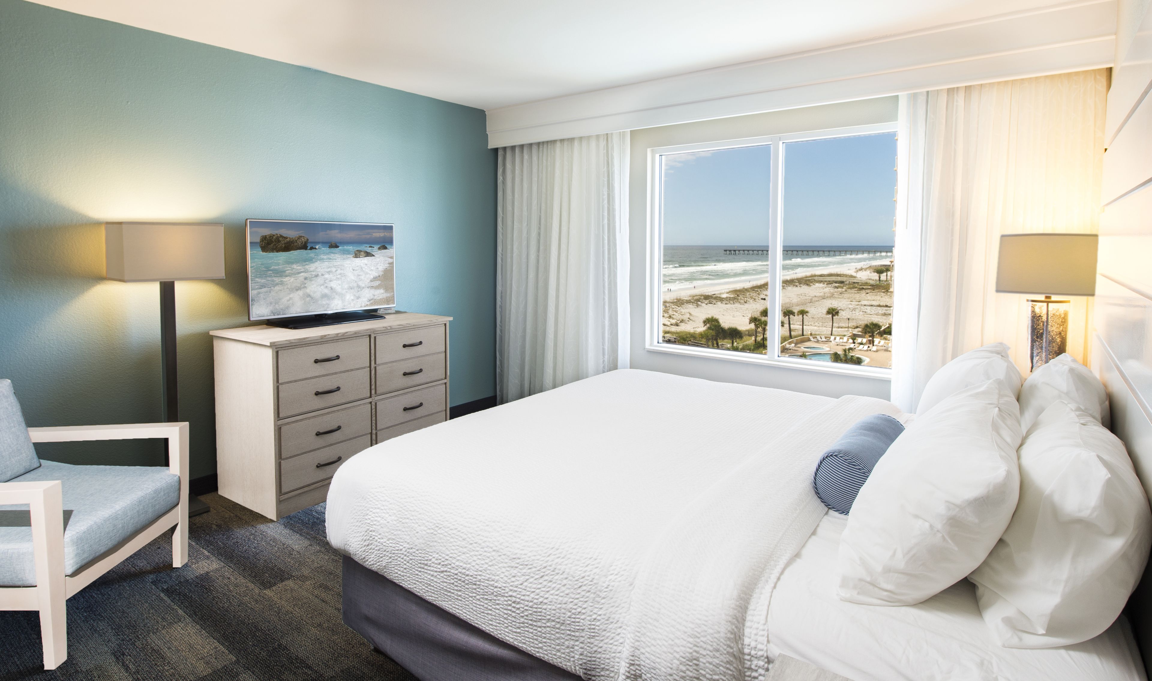 Une chambre d’hôtel avec un lit, un téléviseur posé sur une commode, une chaise et une vue sur la plage et la jetée, visible par la fenêtre.
