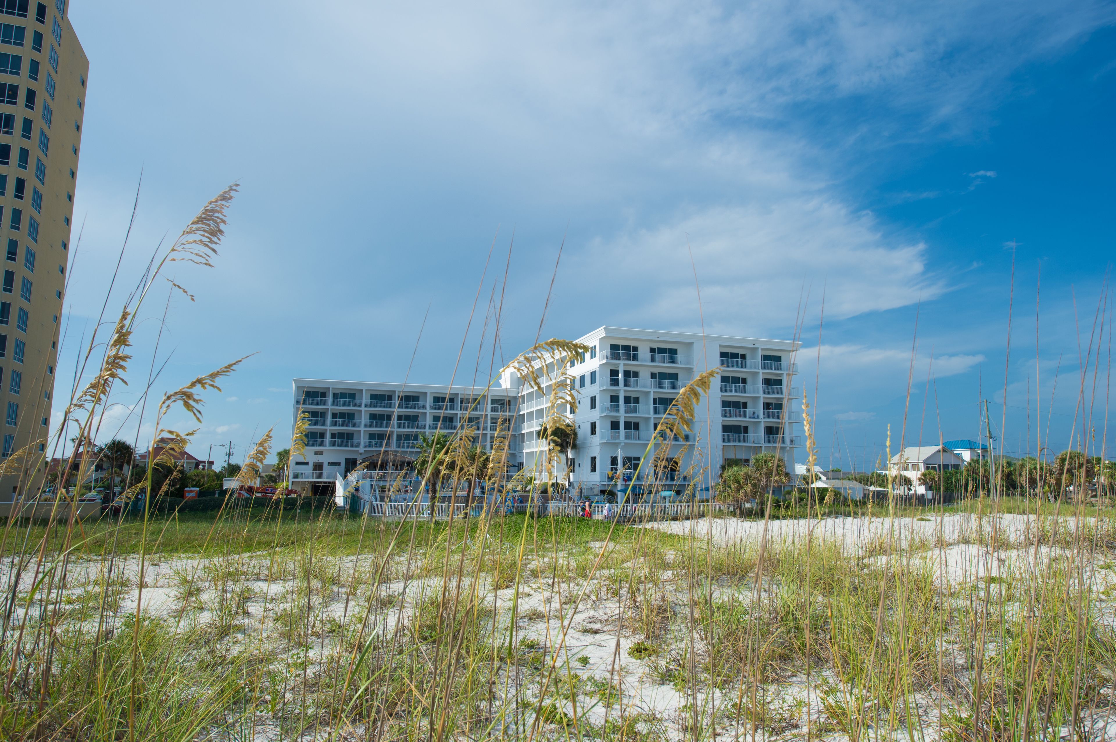 A beachfront hotel with multiple floors, a sandy beach in the foreground, and tall grass.