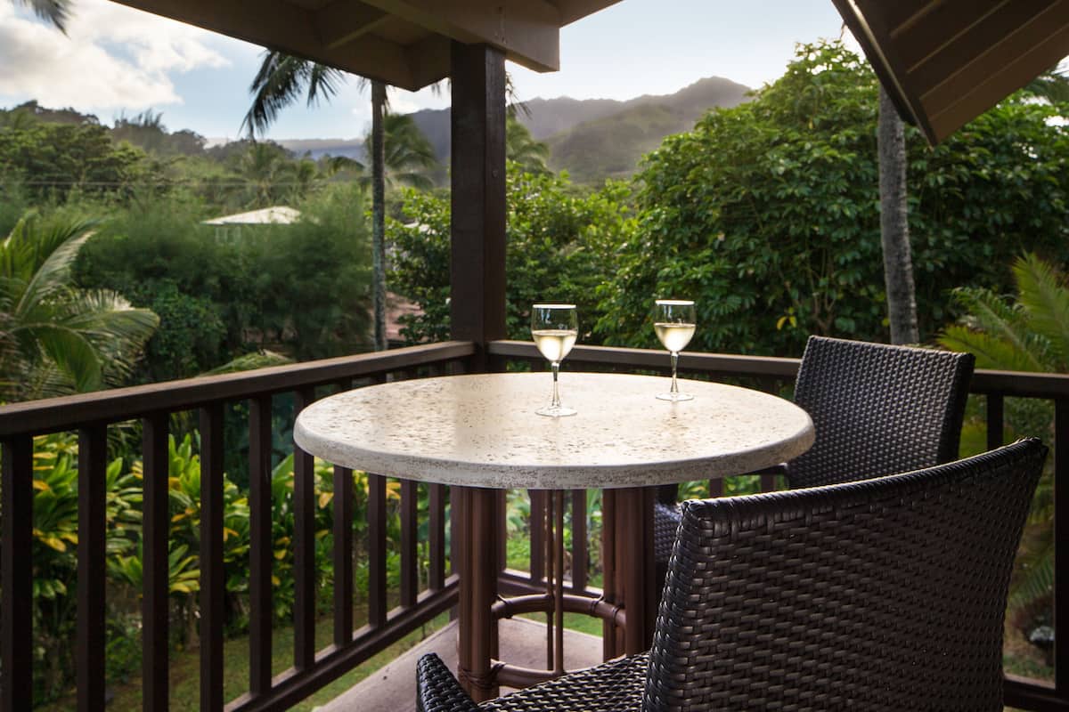A round table with two chairs and two glasses of a beverage, set on a balcony with a view of lush greenery and mountains.