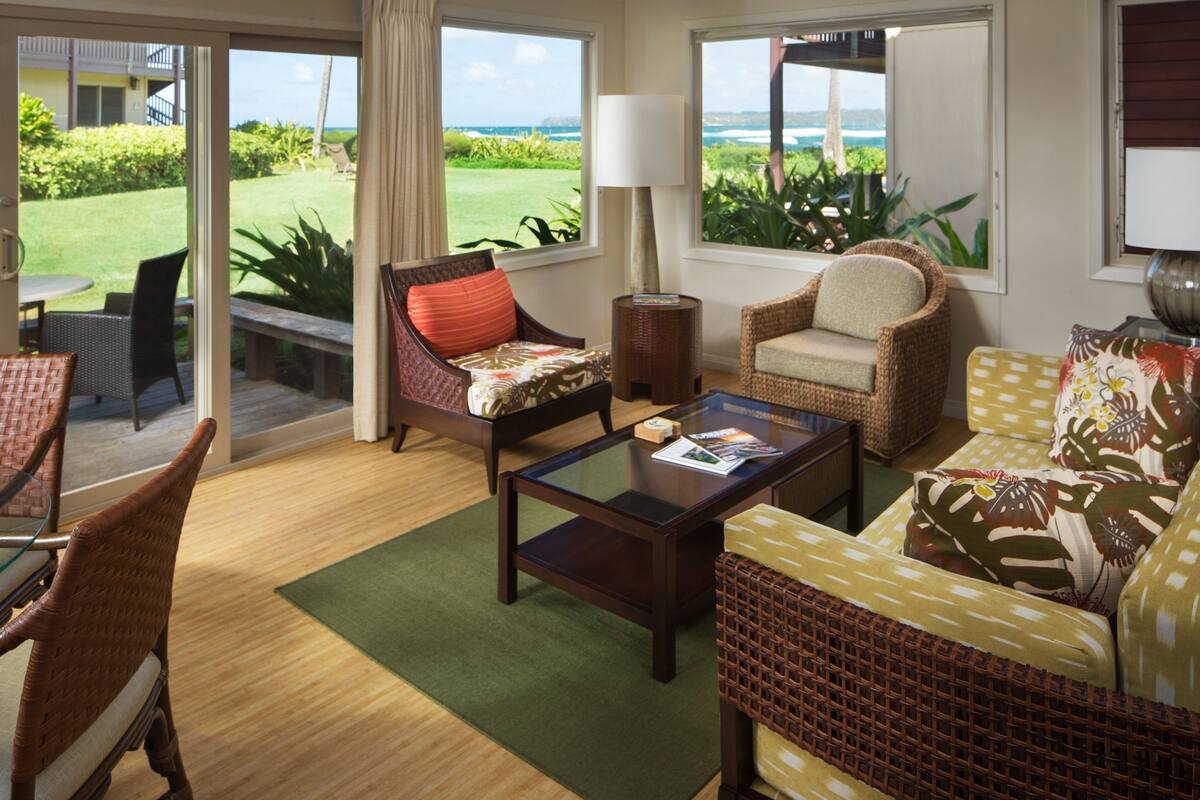 A spacious living room with wicker furniture, a glass coffee table, and a view of a lush garden and ocean through large windows.