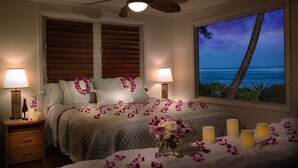 Une chambre à coucher avec un lit, deux lampes de chevet, une table de nuit, un vase de fleurs et une fenêtre donnant sur l’océan.