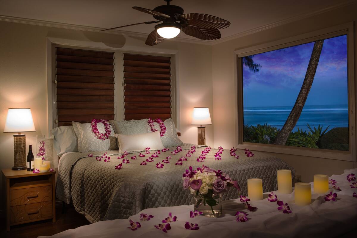 A bedroom with a bed, two bedside lamps, a nightstand, a vase with flowers, and a window with a view of the ocean.