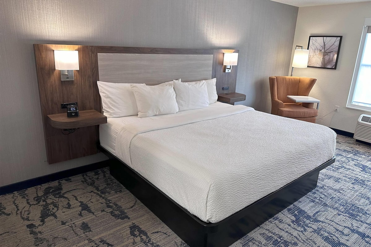 A hotel room with a large bed, two bedside tables, a nightstand, a chair, and a wall-mounted lamp.