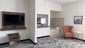 A modern hotel room with a flat-screen TV, a small TV, a seating area with a chair, a side table, and a patterned carpet.