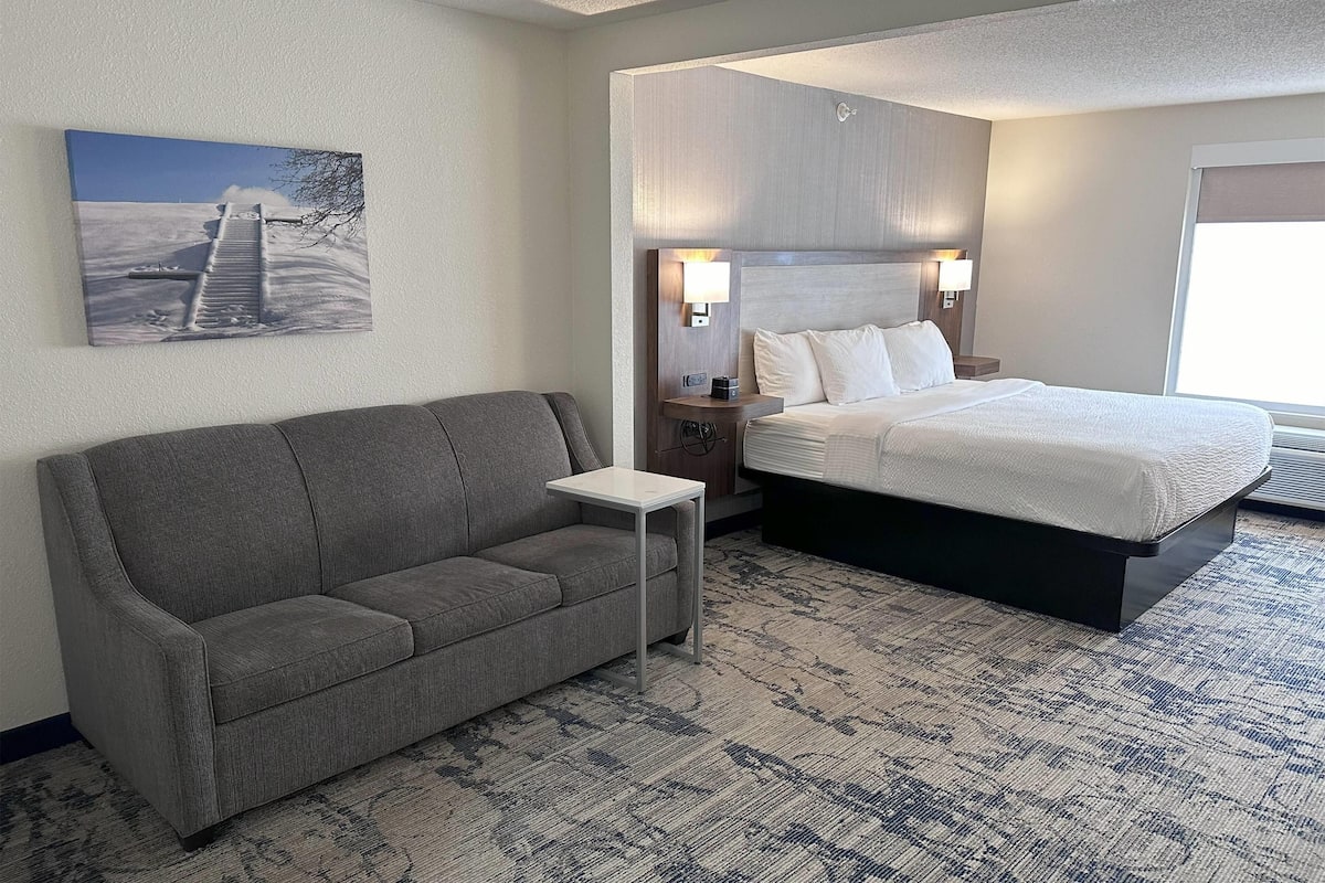A hotel room with a gray sofa, a small white table, a large bed with white bedding, and a painting of a snowy landscape on the wall.