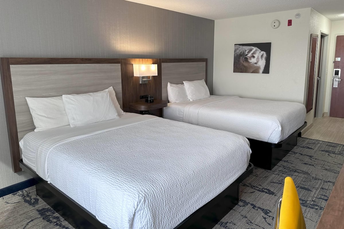 A hotel room with two beds, a nightstand with a lamp, a painting on the wall, and a door to the right.