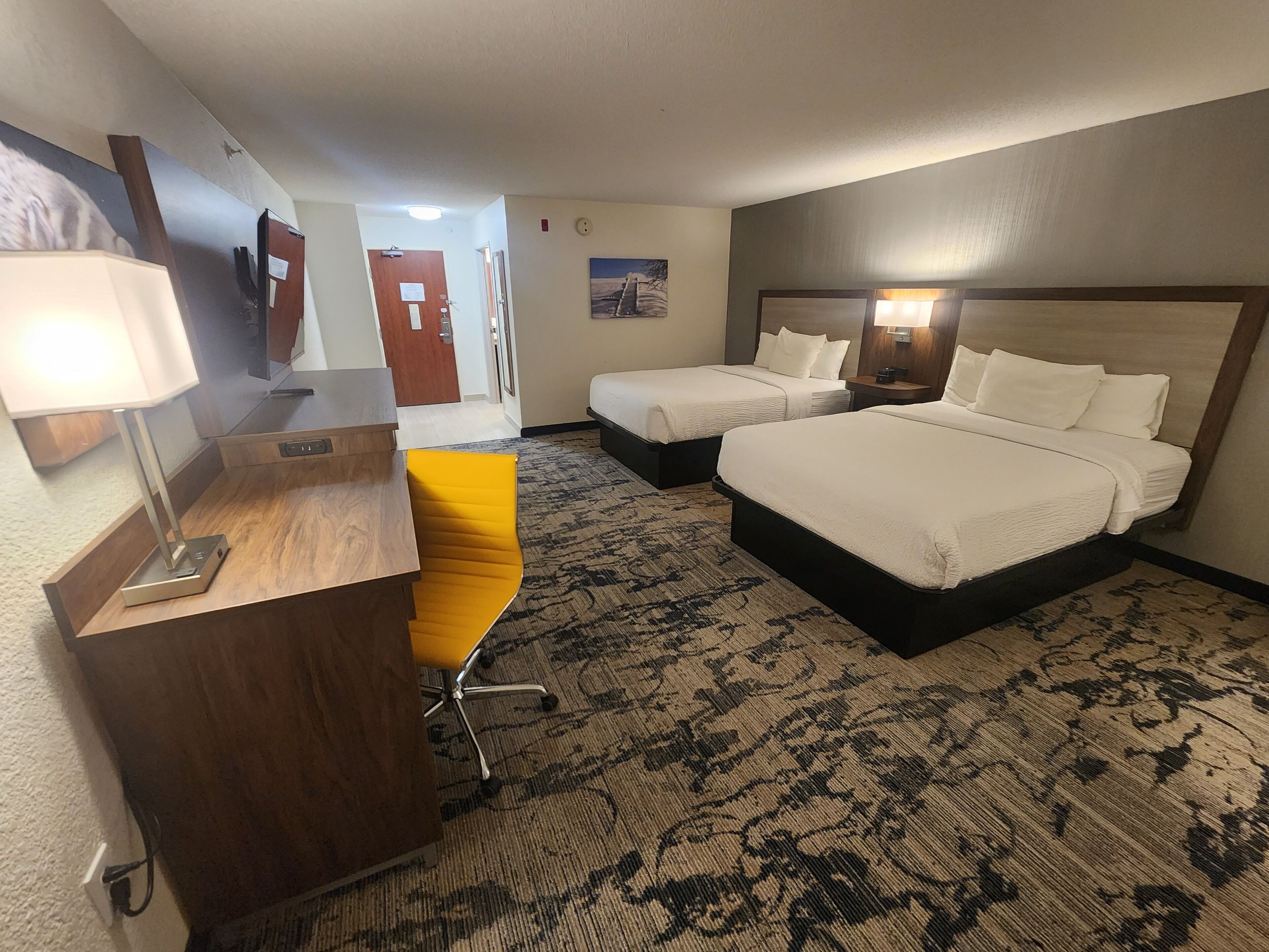 A hotel room with two beds, a desk with a lamp, a chair, and a carpeted floor.
