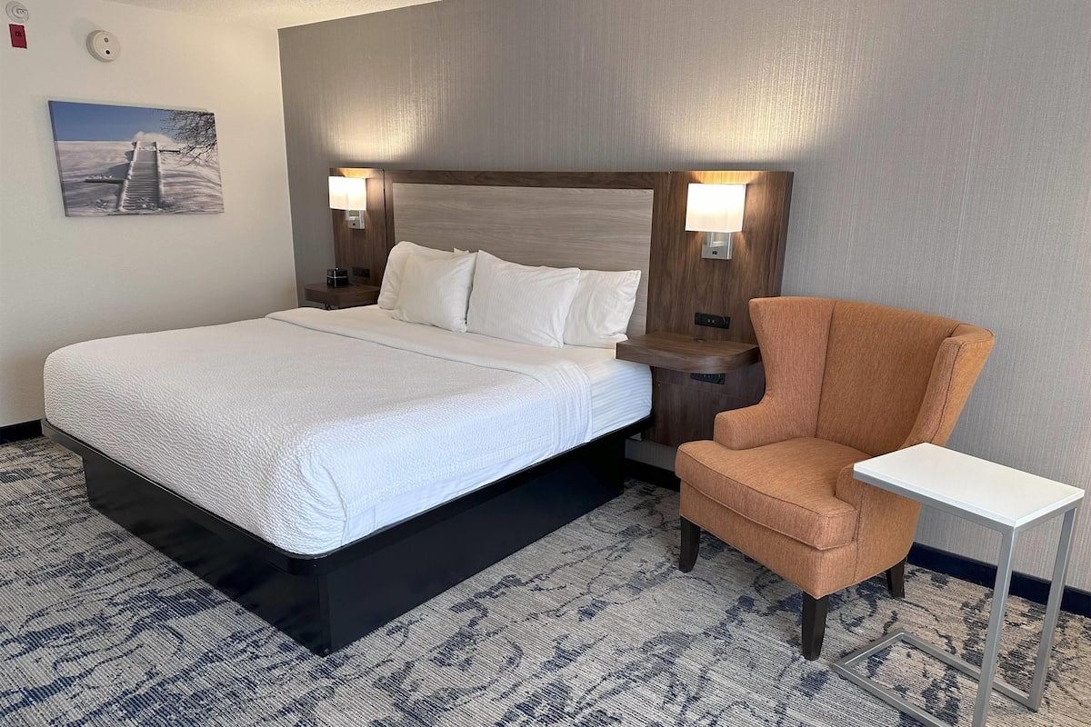 A hotel room with a large bed, a wooden headboard, two bedside lamps, a small armchair, and a side table.