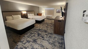 A hotel room with two beds, a desk, a chair, a TV, and a window.