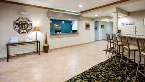 A hotel reception area with a reception desk, a blue wall with the hotel name, a mirror, a lamp, and a patterned rug.
