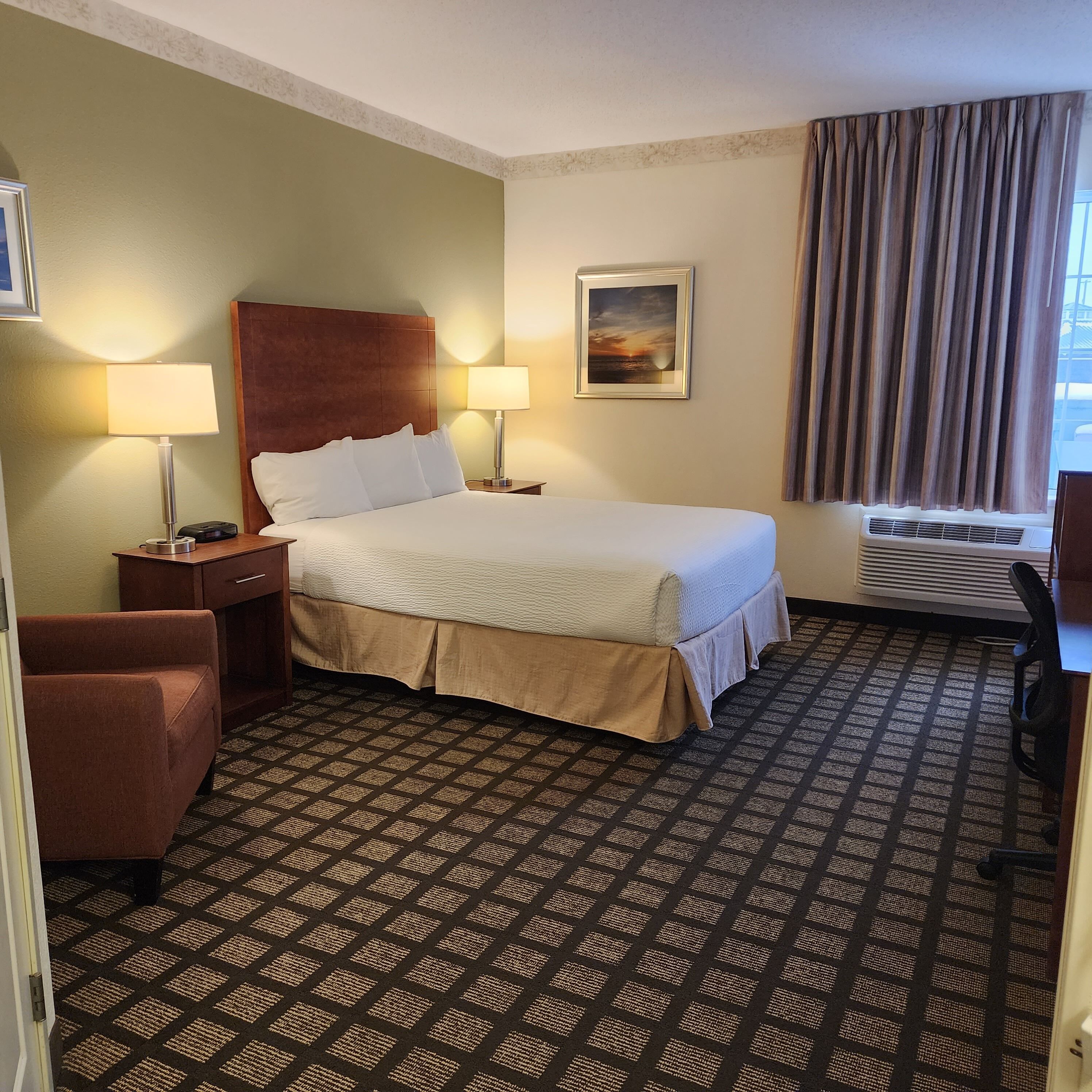 Habitación de hotel con cama, dos lámparas de noche, mesita de noche, una silla, un cuadro, ventana con cortinas y alfombra estampada.