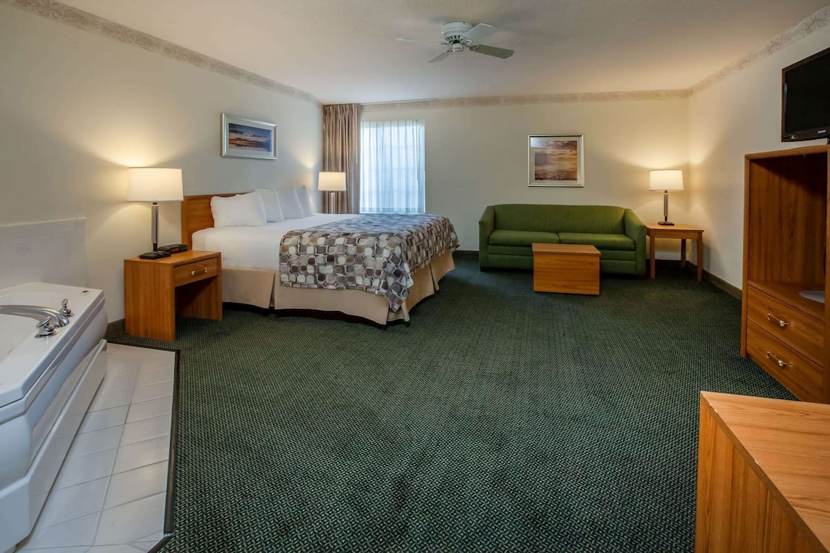 A hotel room with a bed, a green sofa, a wooden cabinet, a TV, and a bathroom with a bathtub.