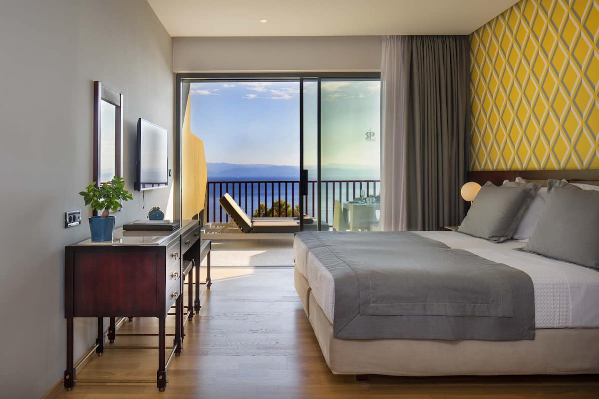 Une chambre d’hôtel moderne avec un grand lit, un bureau agrémenté d’une plante, une télévision et un balcon offrant une vue sur la mer.
