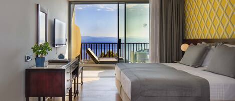 Une chambre d’hôtel moderne avec un grand lit, un bureau agrémenté d’une plante, une télévision et un balcon offrant une vue sur la mer.