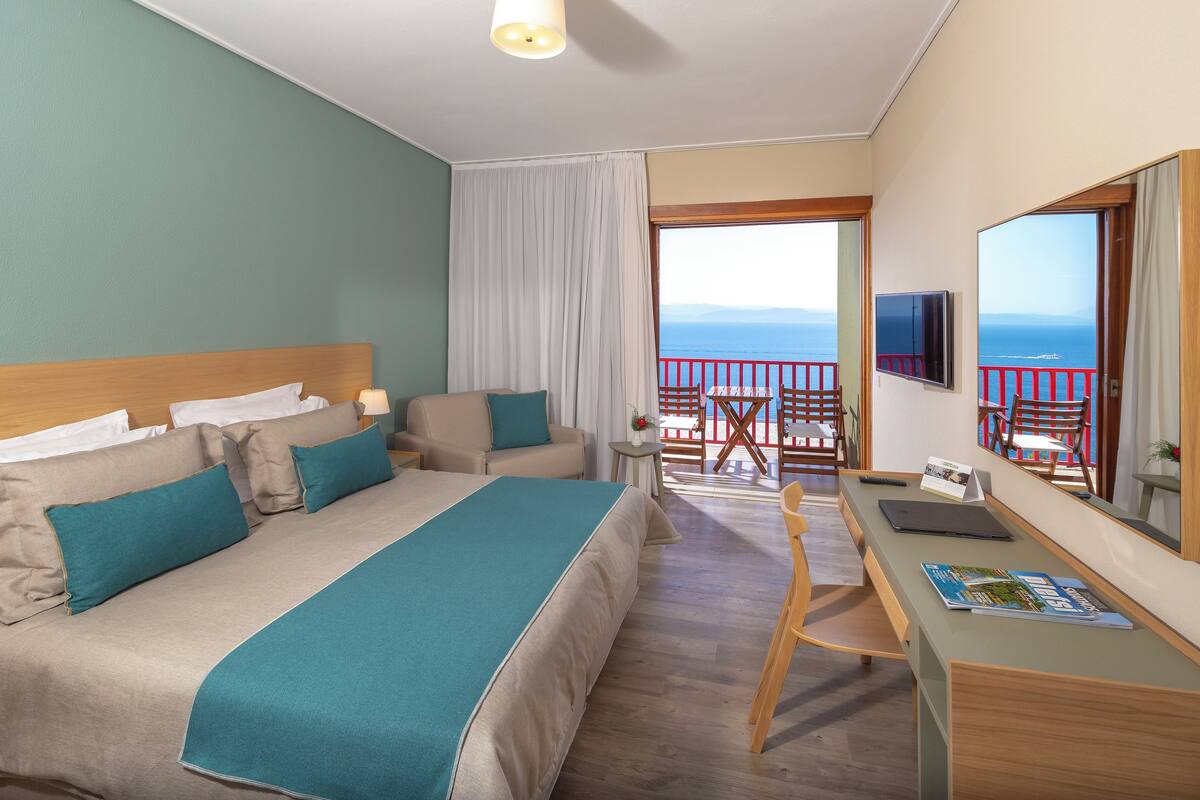 Une chambre d’hôtel équipée d’un lit, d’un bureau, d’une chaise, d’une télévision et d’un balcon avec vue sur la mer.