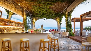 An outdoor bar area with stone flooring, wooden stools, and a view of the sea. - Skiathos Palace Hotel (Skiathos)