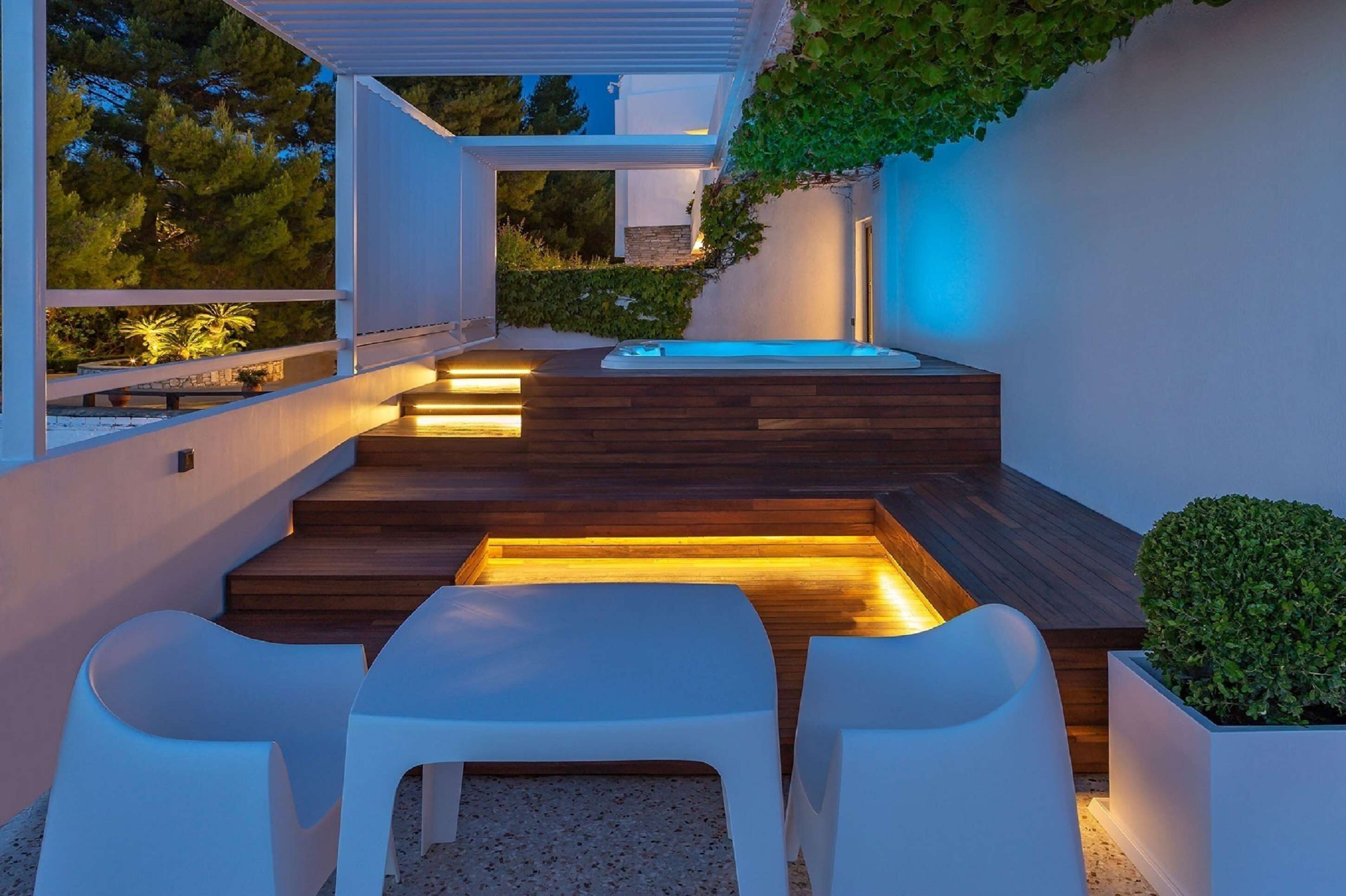 A modern outdoor patio with a wooden deck, white chairs, and a hot tub.