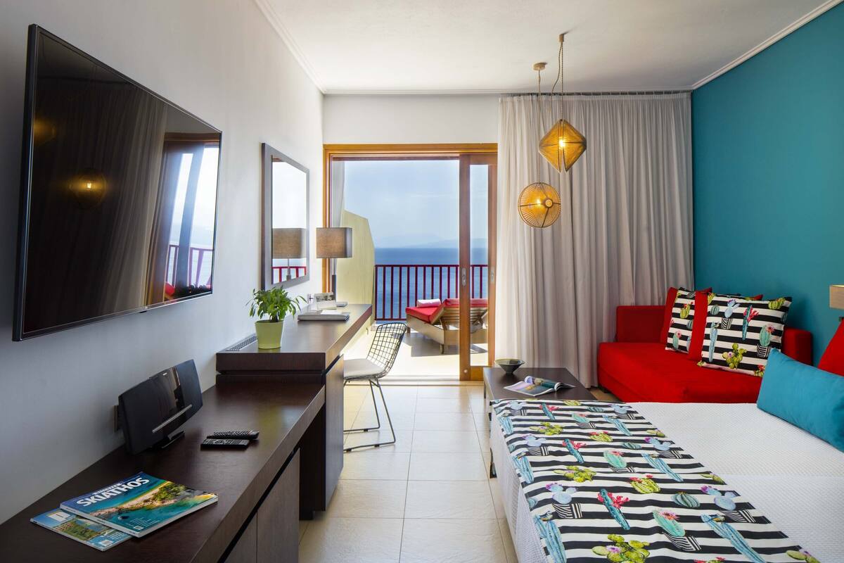 Une chambre d’hôtel avec un lit, un bureau avec une télévision, une chaise, un balcon avec vue et un canapé rouge.