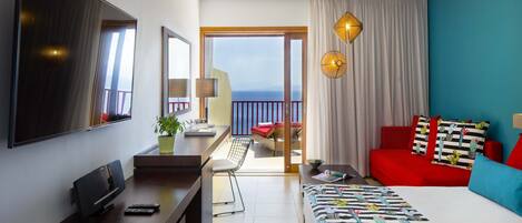 A hotel room with a bed, a desk with a TV, a chair, a balcony with a view, and a red sofa.