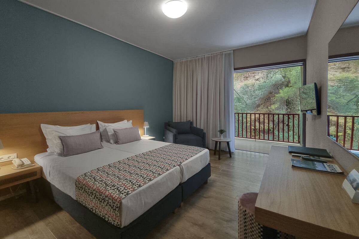 Une chambre d’hôtel avec un grand lit, un bureau, une chaise, une télévision et un balcon donnant sur la verdure.