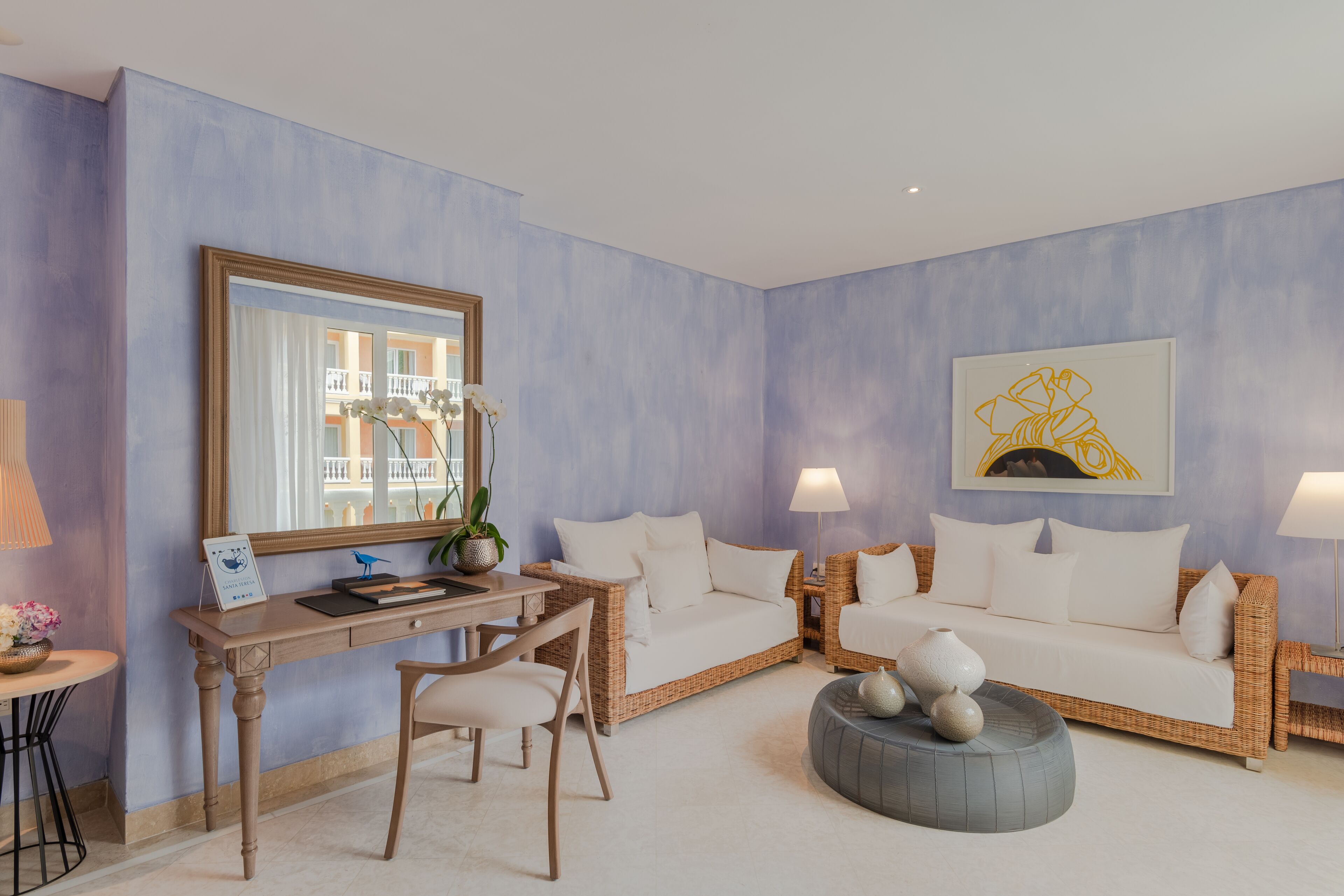 Een kamer met een bureau, twee witte banken, een rond grijze poef, een grote spiegel en een lijst met kunstwerk aan de muur.